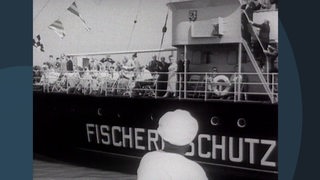 Ein schwarz-weiß Bild, welches Personen auf einem Schiff zeigt. Auf dem Schiff steht die Aufschrift "Fischerschutz".