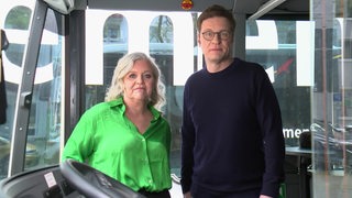 Eine blonde Frau mit einer grünen Bluse steht neben einem Mann mit Brille und dunkelblauem Pullover im Eingangsbereich eines Busses.