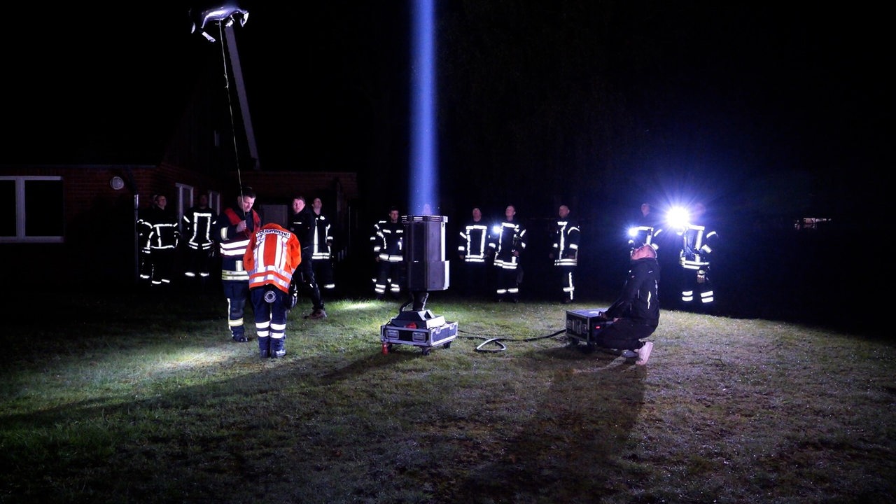 Menschen in Feuerwehrkleidung stehen nachts um einen Scheinwerfer, der einen Lichtkegel nach oben strahlt.
